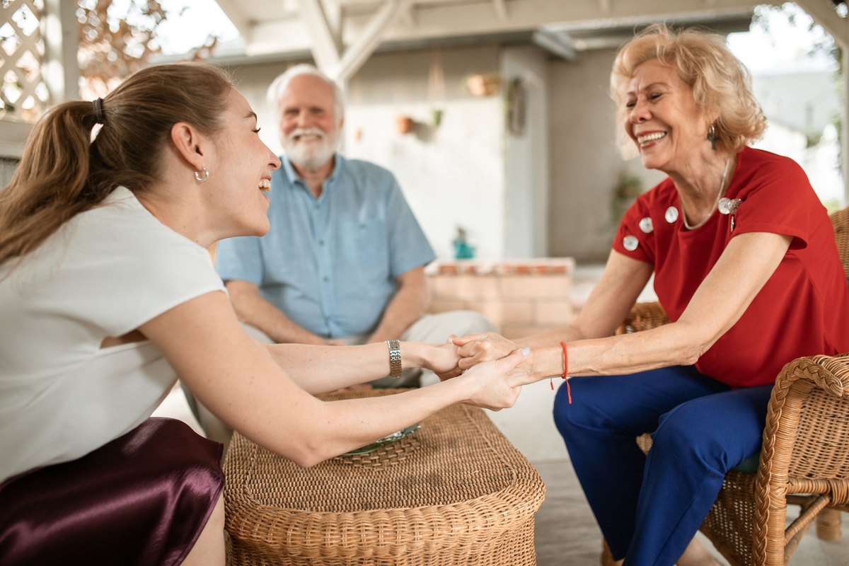 Young woman holding the hands of a laughing older woman beside a smiling older man on a porch