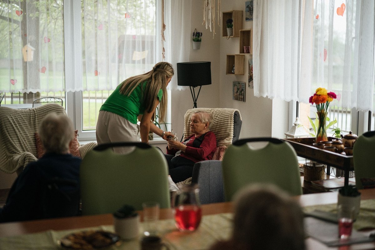 Caregiver in a green top leaning over to hold the hands of an elderly woman in an armchair