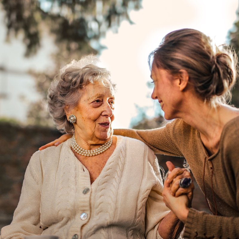 Smiling older woman in a cream cardigan and pearls talking with a younger woman who holds her hand and has an arm around her