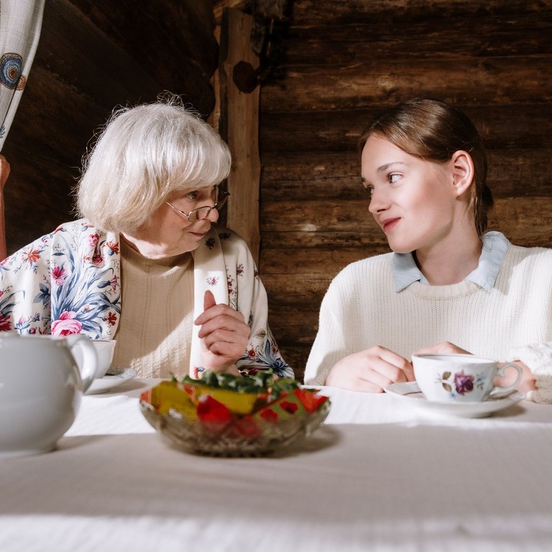 Older woman and younger woman sharing tea at a table with a teapot and fruit bowl